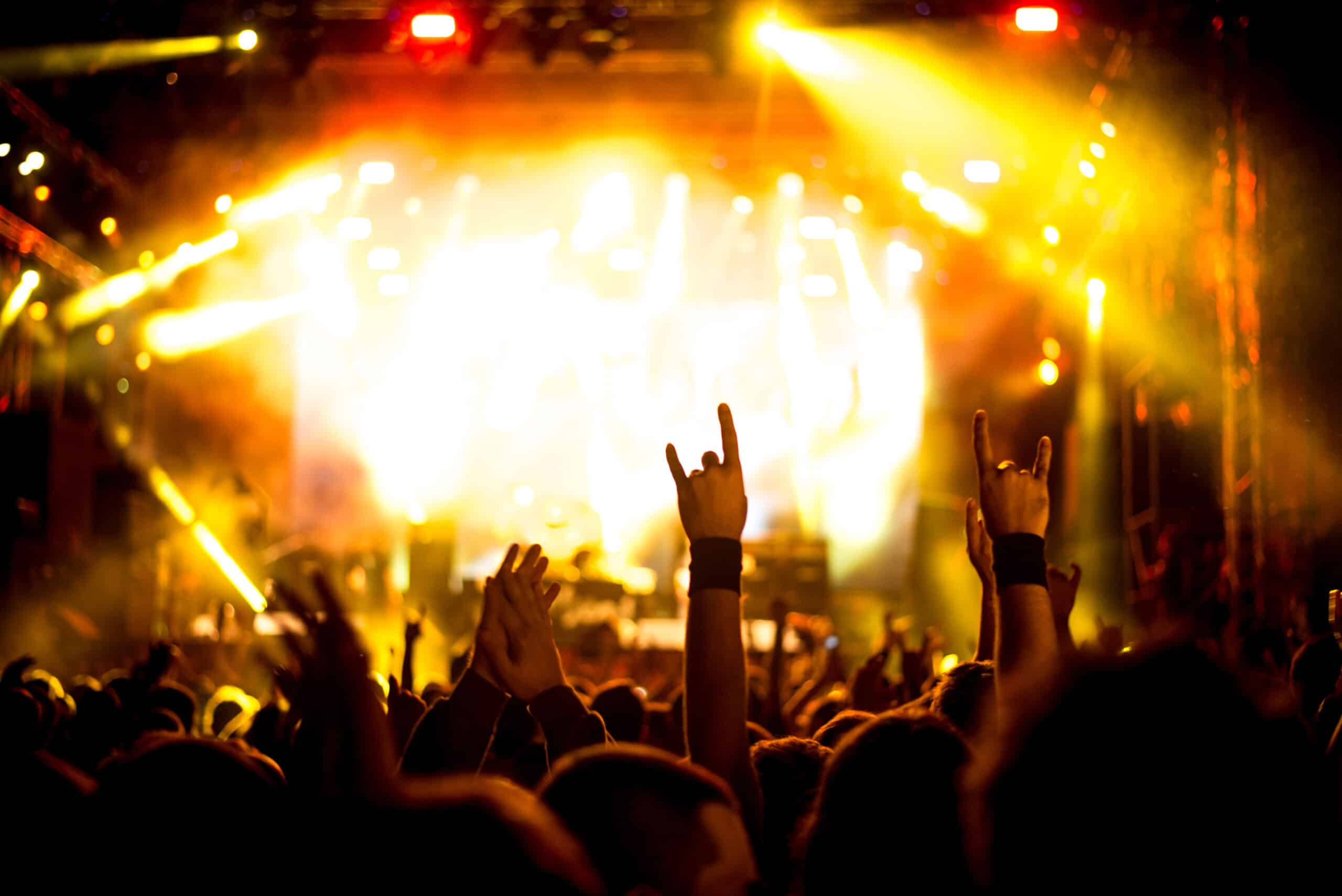 Crowd at a concert raising hands and making rock-and-roll gestures, illuminated by bright yellow stage lights.