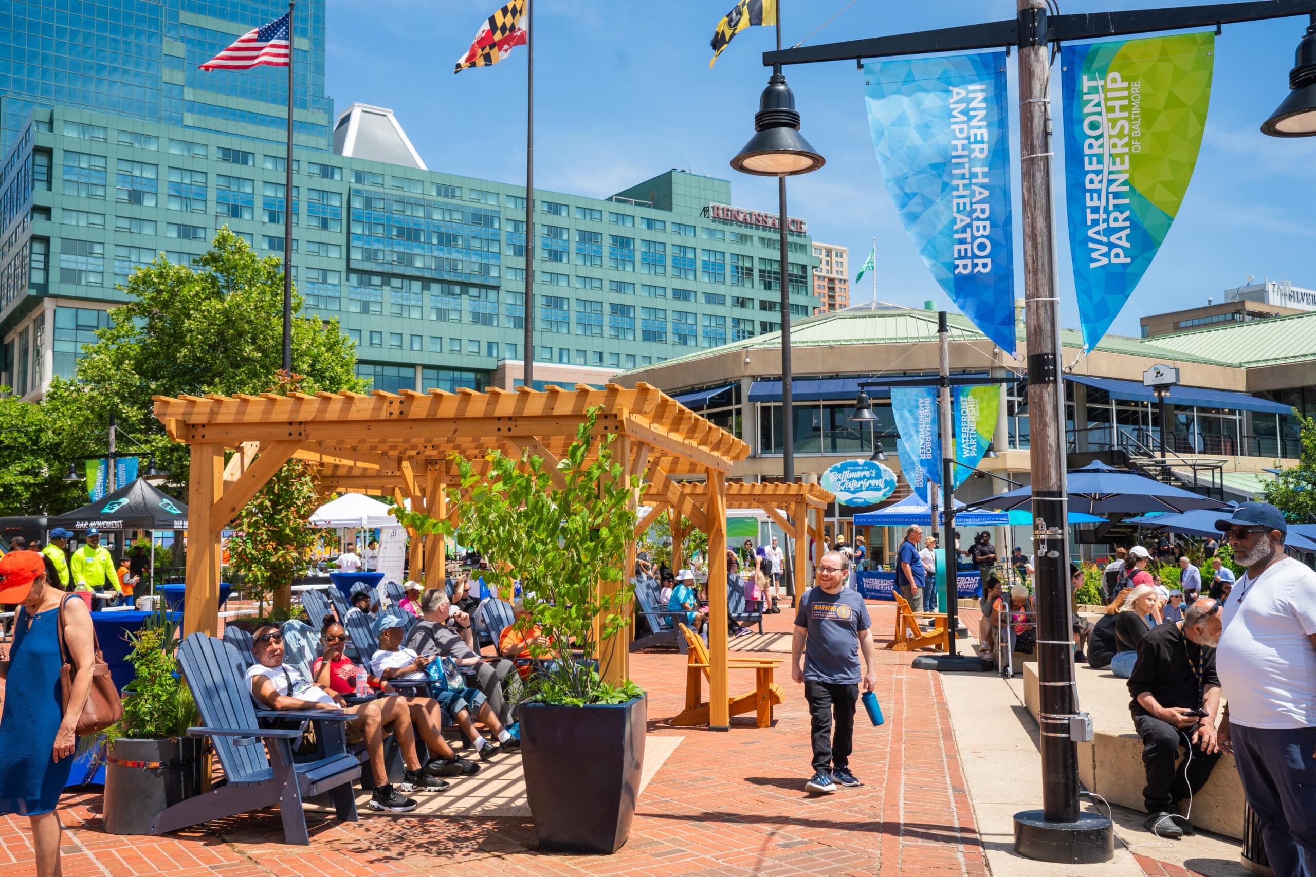 People sit and walk around a shaded seating area in an urban plaza with tall buildings, banners, and flags in the background on a sunny day.