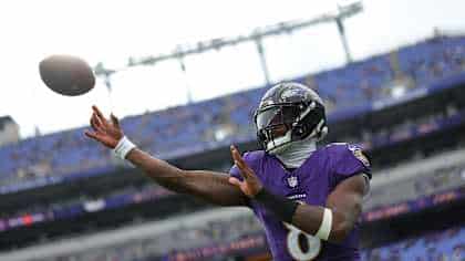 A football player in a purple Baltimore Ravens uniform throws a football on a stadium field with empty blue seats in the background.
