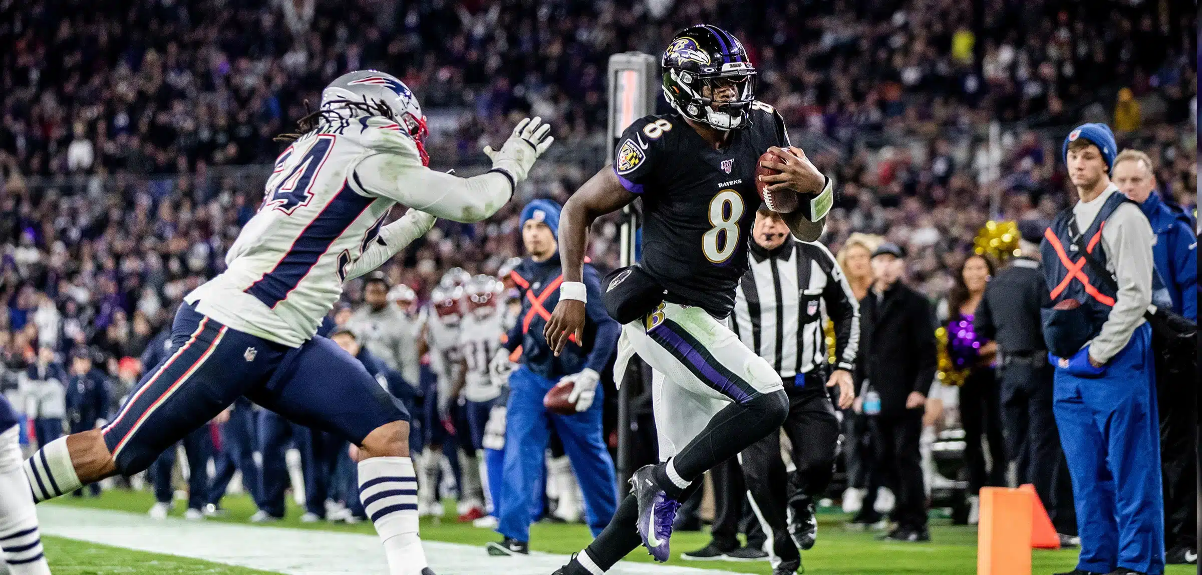 A Baltimore Ravens player runs with the football while being chased by a New England Patriots defender during an NFL game.