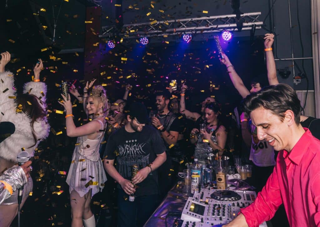 A DJ in a pink shirt performs while people dance, celebrate, and throw confetti in a lively, crowded club with colorful lights.