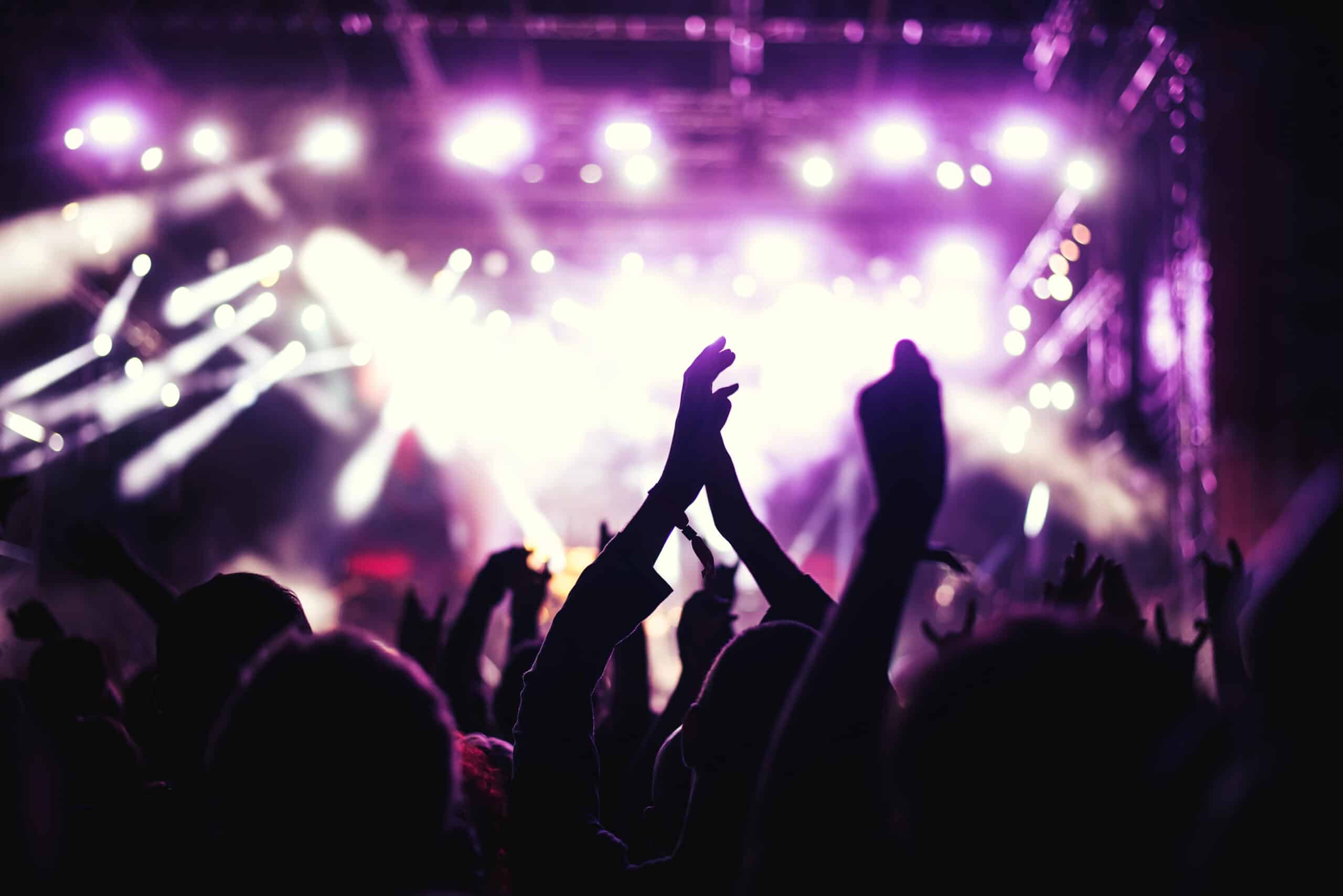 People in a crowd clap and raise their hands at a concert with bright purple and white stage lights in the background.